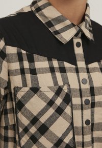 Skjorta med krage i svart och beige rutigt mönster; har svarta axelpaneler, knappar och en bröstficka med matchande rutigt mönster.