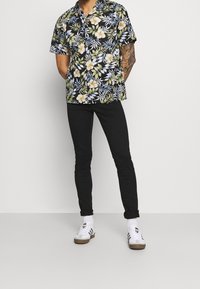 Korte mouwen, zwart bloesemshirt met groene bladeren en gele bloemen, gecombineerd met slanke zwarte broek en witte sneakers met bruine accenten.