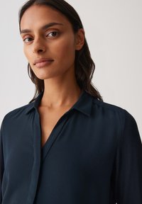 Marineblauwe blouse met een uitgesneden kraag, lange mouwen en een gladde textuur. Kenmerkt zich door een knooploos ontwerp en een subtiele drape.