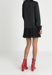 Blusa nera a maniche lunghe con fiocco al collo, abbinata a una gonna nera e stivaletti alla caviglia in pelle laccata rossa brillante con tacco quadrato.