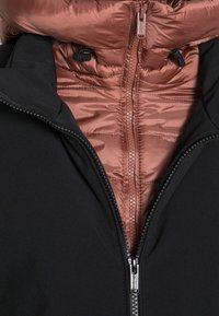 Chaqueta negra con un interior acolchado color rosa dorado. Cuenta con cierres dobles, cordón elástico y cuello forrado. Textura exterior suave.