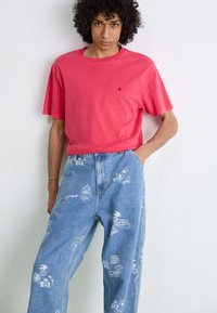 Camiseta roja de cuello redondo con mangas cortas, combinada con jeans de denim azul claro que presentan varios estampados gráficos blancos y un corte relajado.