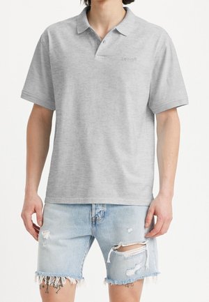 Mand iført en lysegrå kortærmet poloshirt og lyseblå revnede denimshorts, står med armene afslappet ned langs siderne.