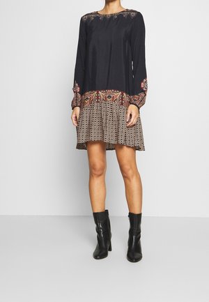 Schwarzes langärmliges Kleid mit besticktem Ausschnitt und gemusterter Rockpartie in erdigen Farben, kombiniert mit knielangen schwarzen Stiefeln.