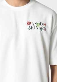 Balta medvilnė marškinėliai su grafiniu spausdinimu "PAS DE MONACO", apsuptu spalvingų gėlių raštų. Trumpi rankoviai ir apvalus kaklo iškirpimas.