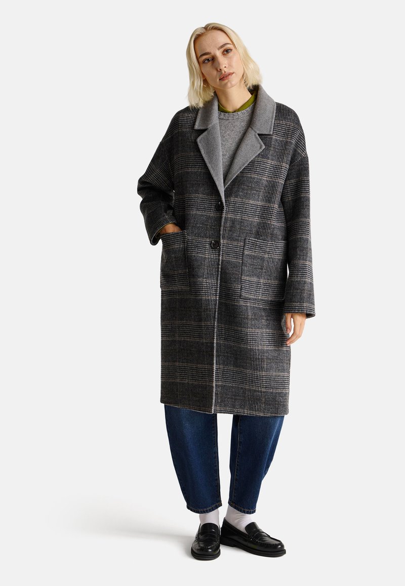 Abrigo de cuadros gris con un gran cuello, dos bolsillos frontales y un corte holgado. Lleva puesto un suéter gris y jeans azules, combinados con zapatos negros.