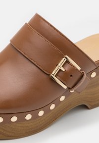 Sabot en cuir marron avec boucle dorée et clous ronds dorés sur semelle plateforme en bois, montrant des coutures détaillées et une texture.