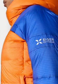 Jachetă izolantă colorată cu secțiuni portocalii și albastre, având un model în grilă, guler înalt și logo-ul EIGER EXTREME pe mânecă.