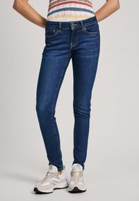 Slim fit mörkblå jeans som bärs med beige och vita sneakers samt en färgglad randig ärmlös topp.