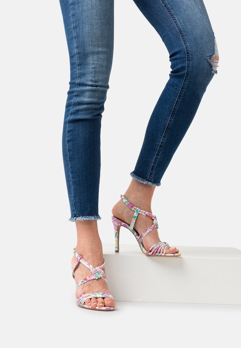 Jambes en jean skinny bleu déchiré portant des sandales à talons hauts fleuries avec des brides autour de la cheville, debout sur un bloc blanc.