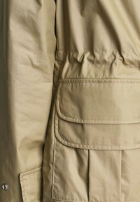 Manche et côté de veste beige avec poche à double rabat et détails cousus en gros plan.