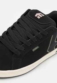 Black sneaker with textured suede upper, flat laces, padded tongue, and subtle green accents. White midsole with branding detail.