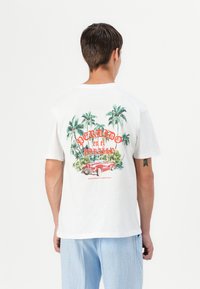 Witte katoenen t-shirt met een kleurrafische afbeelding van palmbomen en een klassieke rode auto op de achterkant, met tekst in een vet lettertype.