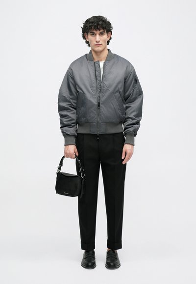 Šedá bomber bunda s žebrovanými manžetami a lemováním, kombinovaná s černými oblekovými kalhotami a černou taškou. Hladká látka, minimalistický design.