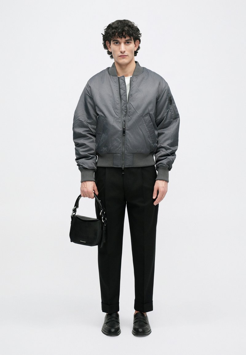 Veste bomber grise avec des poignets et un ourlet côtelés, associée à un pantalon noir ajusté et un sac noir. Tissu lisse, design minimaliste.