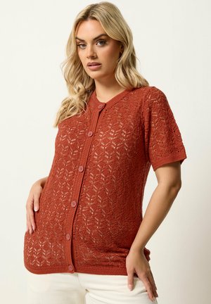 Donna che indossa un cardigan corto a maniche corte color ruggine, lavorato a maglia con bottoni e motivo in pizzo, pantaloni bianchi, posando su sfondo neutro.