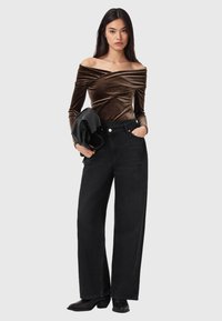 Brun fløjls off-shoulder top kombineret med sort, højtaljet viddeben jeans. Modellen holder en sort jakke med en glat tekstur.