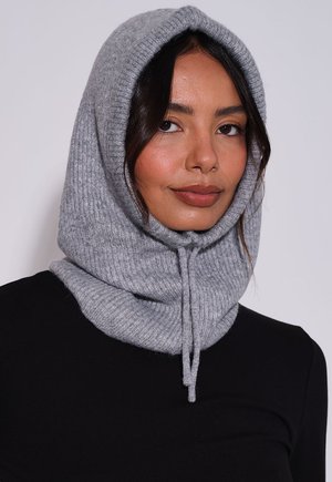 Vrouw draagt een grijze gebreide capuchonsjaal die haar hoofd en nek bedekt, gecombineerd met een zwarte top tegen een effen lichte achtergrond.