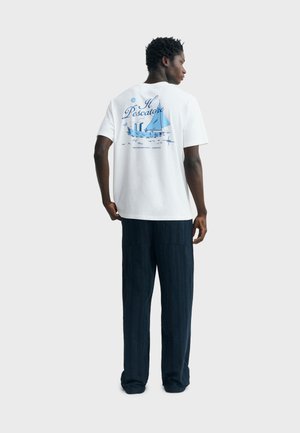 Mann trägt weißes T-Shirt mit blauem Segelboot und dem Text "Il Pescatore" auf der Rückseite, kombiniert mit lockeren dunkelblauen Hosen, steht vor hellem Hintergrund.