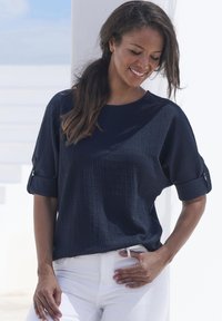 Top navy a maniche corte con un motivo texture, polsini piegati con dettagli a bottoni, abbinato a pantaloni bianchi.