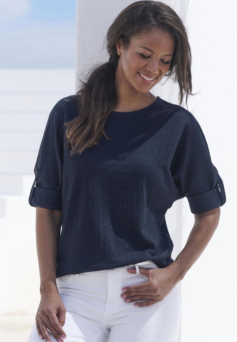 Top navy a maniche corte con un motivo texture, polsini piegati con dettagli a bottoni, abbinato a pantaloni bianchi.