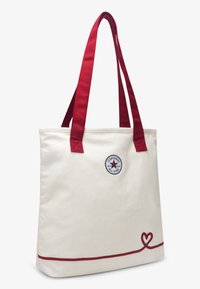 Μπεζ τσάντα ώμου από καμβά με κόκκινους ιμάντες και κόκκινη διακοσμητική γραμμή. Διαθέτει κυκλικό λογότυπο με αστέρι στο κέντρο. Μεγάλος και ορθογώνιος σχεδιασμός.