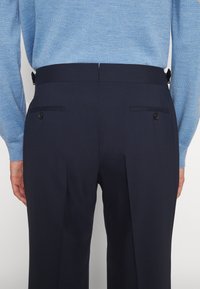 Vue de dos d'une personne portant un pantalon noir ajusté avec des poches arrière boutonnées et un pull bleu clair rentré.