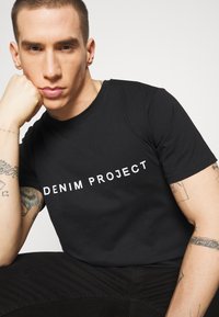 Juodas trumpas rankovėmis marškinėliai su užrašu "DENIM PROJECT" balta spalva, pagaminti iš medvilnės su lygiu paviršiumi, papildyti matomais rankų tetovėjimais.