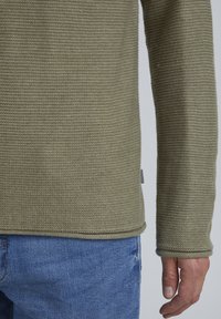 Strickpullover mit langen Ärmeln in olivgrün. Verfügt über eine gerippte Textur und einen geraden Saum. Kombiniert mit hellblauen Denim-Jeans.