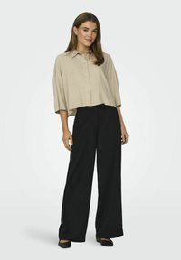 Beige kortærmet skjorte med knapper og krave, parret med sorte vide bukser og flade sorte sko. Stoffet virker glat og let.