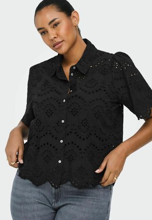 Mujer con una camisa negra de manga corta con estampado de ojales y botones, con dobladillo festoneado y vaqueros azul claro, posando con las manos en las caderas.