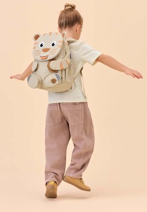 Kind mit Haarknoten, trägt beiges Hemd und Hose, balanciert mit ausgestreckten Armen und trägt einen beigen tigerförmigen Rucksack.
