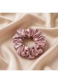 Roze satijnen scrunchie versierd met kleine kraaltjes ligt op glad beige stof.