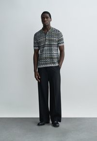 Mees seisab ühevärvilise tausta ees, kandes mustritatud polo särki, musti laiade säärtega pükse ja musti kingi, üks käsi taskus.