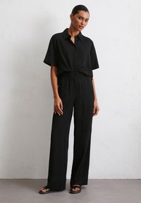 Schwarzes kurzärmliges Hemd mit Knopfleiste, kombiniert mit hochgeschnittenen, weiten Hosen. Glatter Stoff, lockere Passform und elastischer Bund. Sandalen runden das Outfit ab.