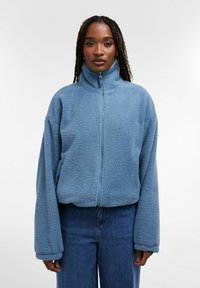 Femme aux longs cheveux tressés portant une veste polaire zippée bleu clair et un jean bleu, debout devant un fond blanc uni.