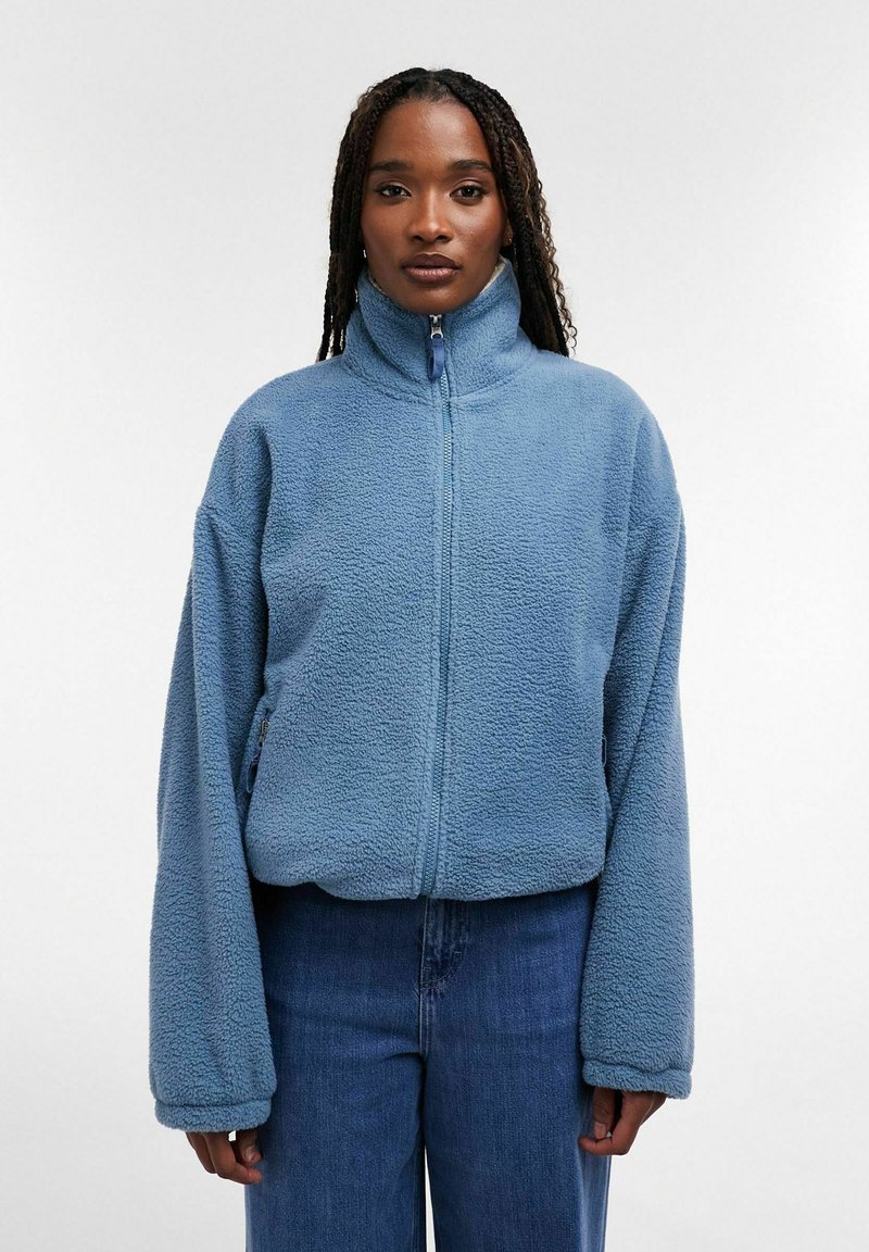 Femme aux longs cheveux tressés portant une veste polaire zippée bleu clair et un jean bleu, debout devant un fond blanc uni.