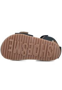 Bruine rubberen buitenzool met een gestructureerde grip, voorzien van het woord "SHOESME" in verhoogde letters. Bandjes van donker materiaal bevestigen zich veilig.