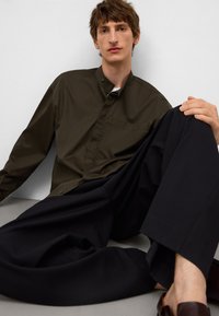 Olijfgroene button-up shirt met mandarin kraag, gecombineerd met ruime zwarte broek, zittend op een grijze ondergrond.