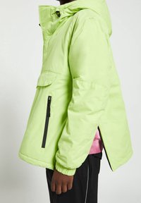 Persona con una chaqueta con capucha de color verde claro, con un bolsillo con cremallera negra y puños elásticos, combinada con pantalones negros y una camisa rosa visible.
