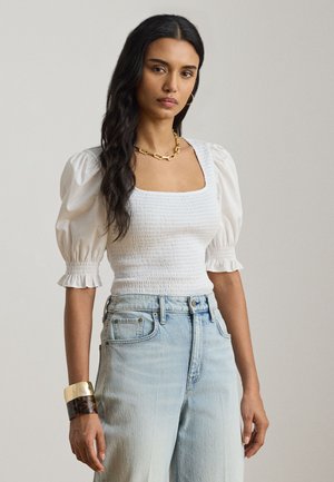 Γυναίκα με μακριά σκούρα μαλλιά που φοράει λευκό τοπ με πούπουλα στα μανίκια και ραφή, ανοιχτό μπλε ψηλόμεσο τζιν, χρυσή αλυσίδα λαιμού και φαρδιά βραχιόλια.