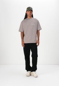 Camiseta gris de gran tamaño con texto blanco, pantalones negros y zapatillas beige. El portador tiene el cabello largo y lleva puesta una gorra de cuadros verde.