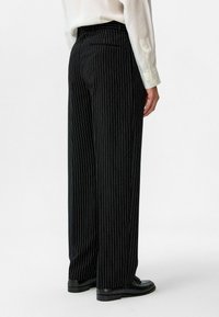Zwarte pinstripebroek met een relaxte pasvorm, voorzien van een hoge taille, zijzakken en een gladde textuur. G gedragen met een lichtgekleurde blouse.