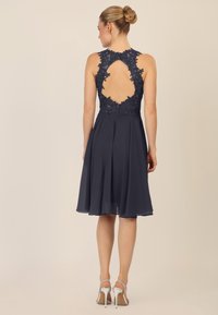 Navyblaues Kleid mit Spitzenverzierungen, offenem Rücken und ausgestelltem Rock. Der Stoff ist leicht und hat eine glatte Textur. Silberne Absatzsandalen.