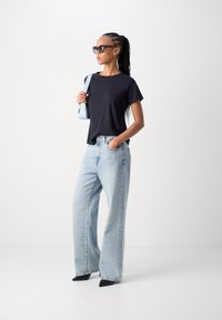 Zwarte crew-neck t-shirt, lichtblauwe wijde jeans, spitse zwarte hakken en oversized zonnebril. Schoudertas in lichtblauw. Gladde texturen.