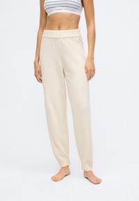 Pantalones de lounge beige de cintura alta, confeccionados en un tejido suave, con un ajuste holgado y puños acanalados en los tobillos. Textura lisa, diseño minimalista.