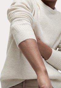 Krämfärgad tröja med en texturerad, stickad mönstring. Ribbstickade manschetter och rundad halsringning. Tygdet verkar mjukt och något elastiskt.