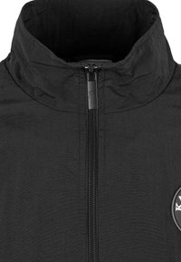 Svart zip-up-jacka med hög krage. Har en glansig dragkedja, en jämn textur och en cirkulär logotypapplikation på vänster sida.