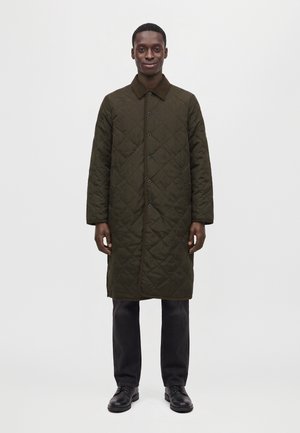 Un homme se tient devant un fond blanc uni et porte un manteau matelassé boutonné, vert olive foncé, un pantalon noir et des chaussures en cuir.