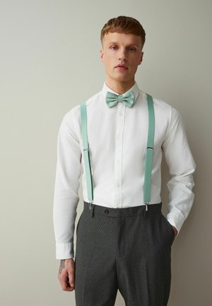 Mladý muž v bielej košeli, mäta zeleným motýlikom a zodpovedajúcimi trakovými nohavicami tmavošedej farby, stojaci pred jednofarebným svetlým pozadím.